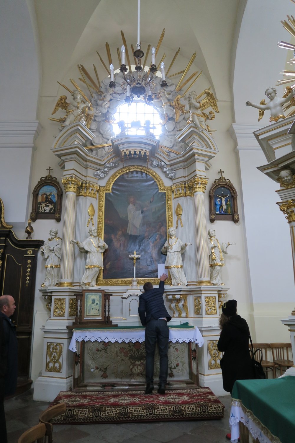 Wnętrze kościoła z ozdobnym barokowym ołtarzem: centralny duży obraz w złotej ramie otoczony białymi kolumnami, rzeźbami aniołów i złotymi promieniami pod wysokim oknem oraz żyrandolem, na ołtarzu krzyż i koronkowy obrus, przed nim mężczyzna w kurtce podnosi rękę ku obrazowi, obok stoi kobieta, a po lewej widoczny jest inny mężczyzna.