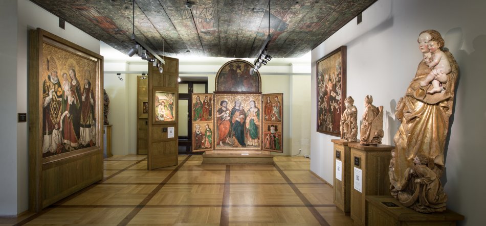 Wnętrze galerii muzealnej z jasną parkietową posadzką i malowanym drewnianym sufitem, centralnie ustawionym dużym tryptykiem malarskim przedstawiającym siedzącą kobietę z dzieckiem i towarzyszące postacie w szatach, po bokach inne obrazy wiszące na ścianach oraz na prawym planie kilka drewnianych rzeźb na postumentach oświetlonych reflektorami.