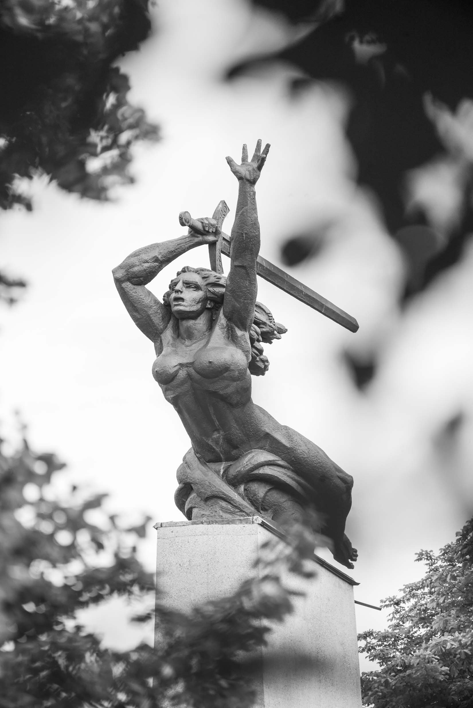 Czarno-białe zdjęcie dużej rzeźby kobiecej postaci ustawionej na wysokim cokole z odsłoniętym torsem, unoszącej oburącz miecz nad głową, w otoczeniu rozmytych liści drzew.
