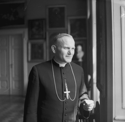 Czarno-białe zdjęcie mężczyzny w sutannie z koloratką i łańcuchem z krzyżem, stojącego przy oknie i spoglądającego przez nie; w tle widoczne obrazy na ścianie, marmurowe popiersie i drzwi.