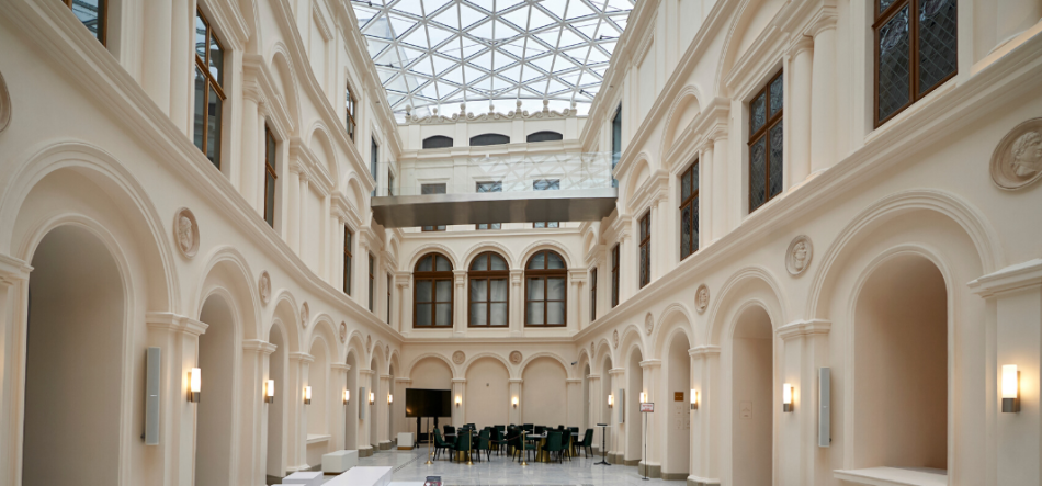Jasne wnętrze atrium z wysokimi kremowymi ścianami i równymi arkadami po obu stronach, dekoracyjnymi medalionami nad łukami, drewnianymi oknami na piętrze, szklaną kratownicową kopułą dachową i zawieszoną przeszkloną kładką, ściennymi kinkietami, białymi ławkami wzdłuż boków oraz stolikami i krzesłami ustawionymi na końcu przestrzeni.