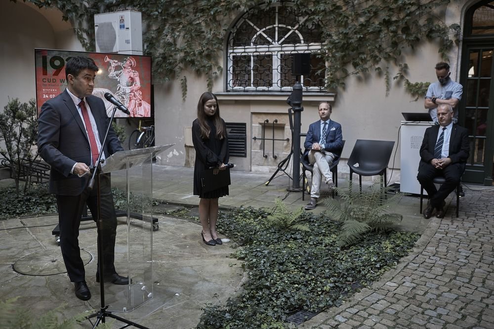 Na kamiennym dziedzińcu mężczyzna w garniturze stoi przy przezroczystym pulpicie i mówi do mikrofonu, obok stoi kobieta w czarnej sukience trzymająca teczkę, po prawej siedzą trzej mężczyźni na krzesłach, a w tle widać okno z metalową kratą, ścianę porośniętą bluszczem oraz ekran z plakatem.