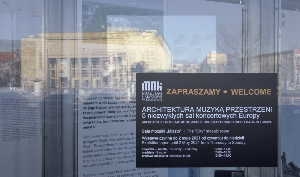 Szyba wejściowa z odbiciem kamiennego budynku, na której przylepiony jest czarny prostokątny plakat Muzeum Narodowego w Krakowie z dużym napisem "ZAPRASZAMY • WELCOME" oraz szczegółowymi informacjami po polsku i angielsku o wystawie "ARCHITEKTURA MUZYKĄ PRZESTRZENI — 5 niezwykłych sal koncertowych Europy" i godzinach otwarcia.