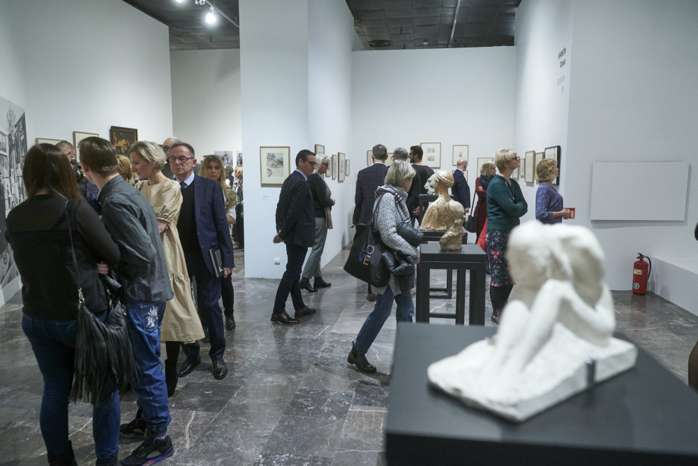 Wnętrze galerii sztuki z białymi ścianami i marmurową podłogą, grupami osób oglądających oprawione rysunki na ścianach, dwiema rzeźbami na czarnych podestach na pierwszym planie, widoczną kolumną pośrodku oraz gaśnicą przy prawej ścianie.