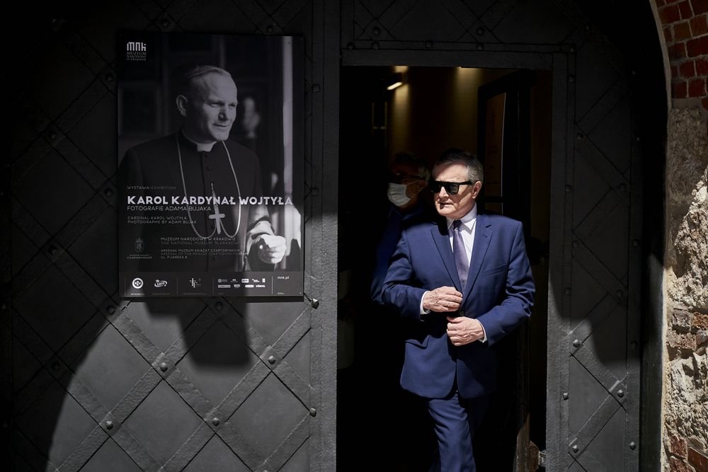 Mężczyzna w granatowym garniturze i ciemnych okularach wychodzi z ciemnego wejścia po prawej, za nim częściowo widoczna osoba w maseczce, a po lewej na metalowych drzwiach wisi czarno-biały plakat z portretem duchownego i dużym białym napisem, całość oświetlona ostrym kontrastowym światłem i cieniem.