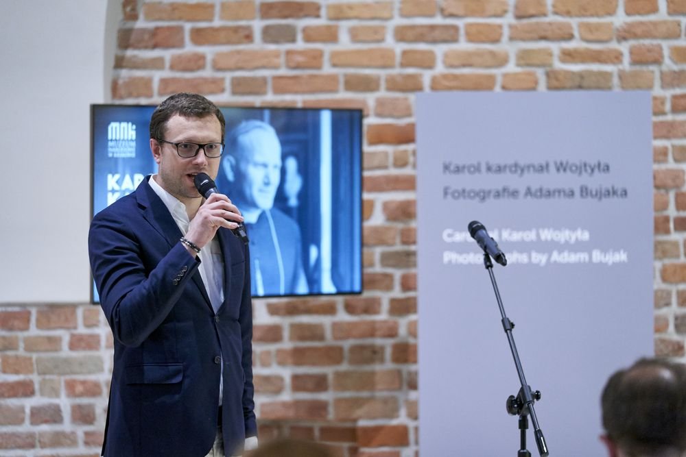Mężczyzna w granatowej marynarce i okularach trzyma mikrofon i przemawia stojąc przed ceglaną ścianą, z lewej za nim ekran z czarno-białym portretem, a z prawej pionowy panel informacyjny i mikrofon na statywie, na pierwszym planie widoczne fragmenty głów słuchaczy.