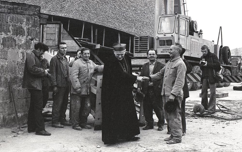 Czarno-białe zdjęcie przedstawiające plac budowy z grupą robotników stojących po lewej i dźwigiem w tle, w centrum mężczyzna w długim ciemnym płaszczu i nakryciu głowy ściska dłoń robotnika trzymającego kask, a po prawej stoi fotograf z aparatem; na pierwszym planie widoczne narzędzia i gruz.