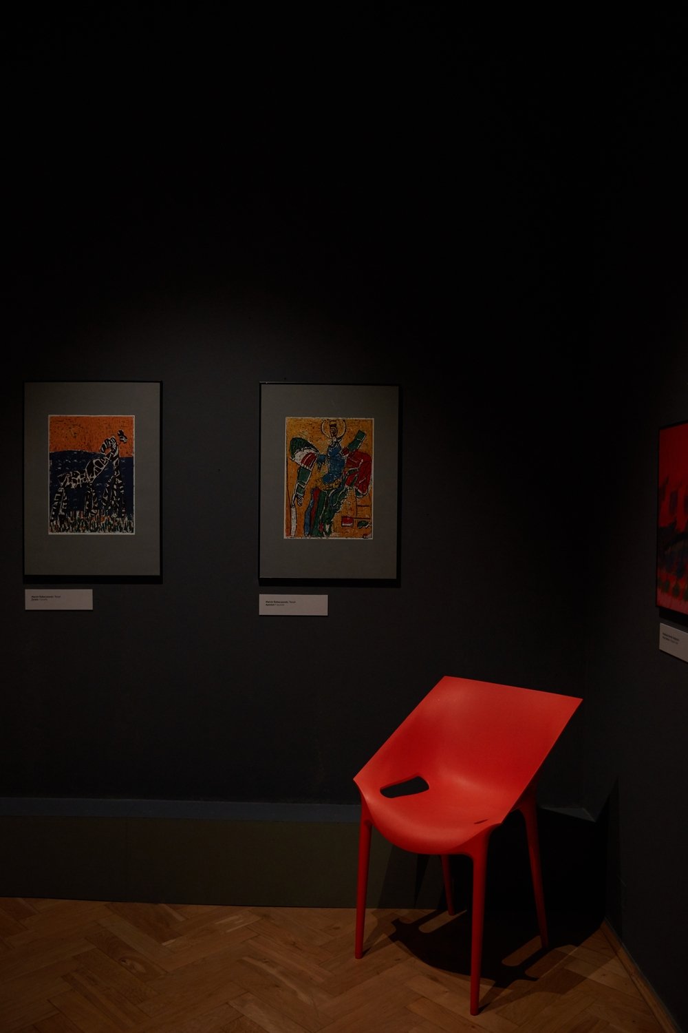 Ciemne wnętrze galerii z dwiema oprawionymi, kolorowymi grafikami z etykietami na ścianie, fragmentem innego obrazu u prawego brzegu oraz jaskrawoczerwonym, nowoczesnym plastikowym krzesłem z otworem w siedzisku ustawionym w kącie na drewnianej podłodze ułożonej w jodełkę.