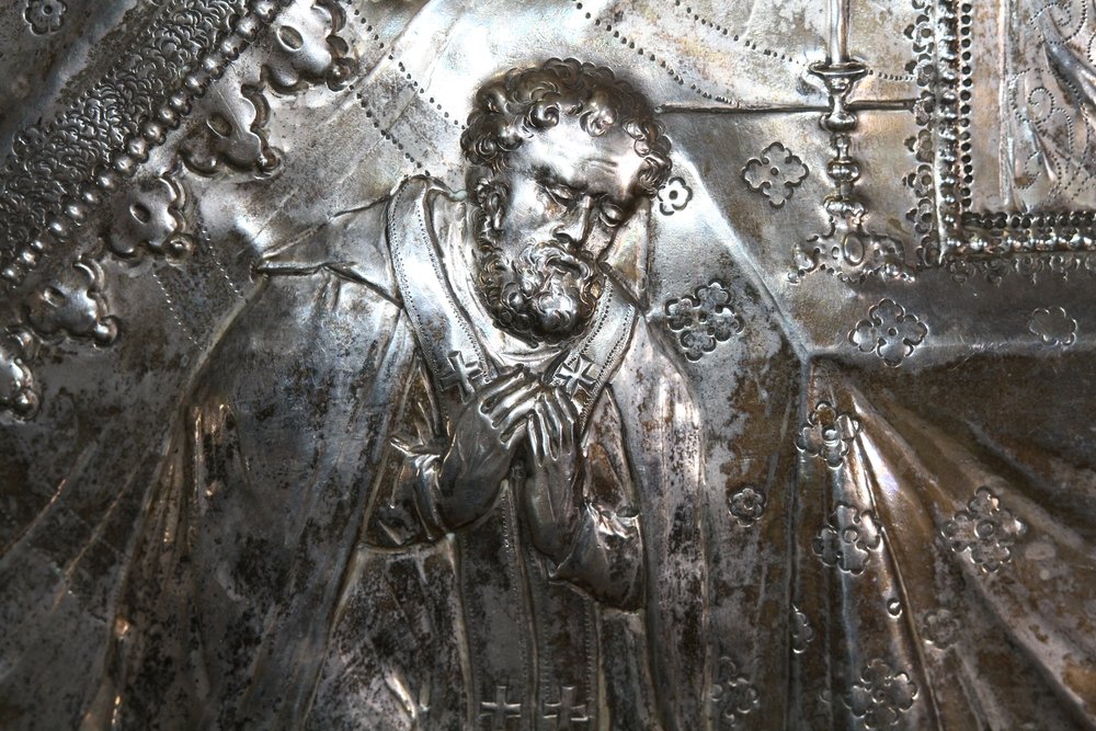 Srebrny tłoczony relief przedstawiający brodatą postać w szatach sakralnych z rękami złożonymi na piersi, z detalami krzyżyków na ornacie, ozdobnym tłem z drobnymi kwiatowymi motywami i świecznikiem po prawej.