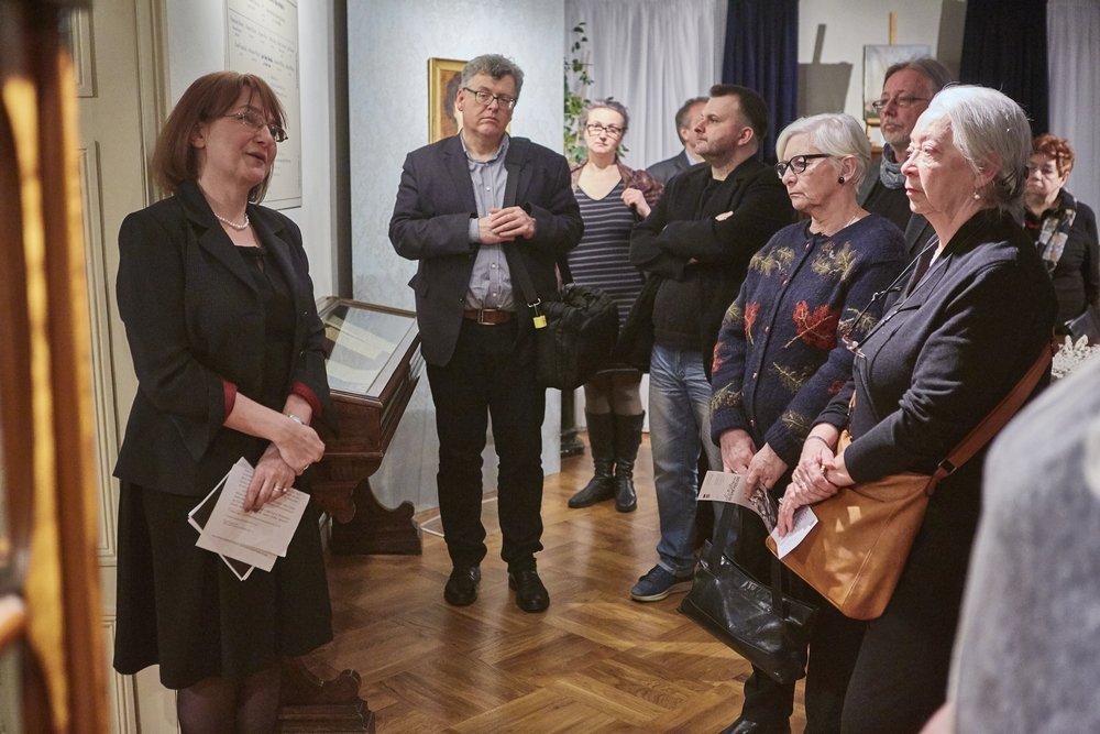 Kobieta po lewej w ciemnym ubraniu trzyma kartki i mówi do grupy dorosłych stojących w półokręgu w pomieszczeniu z drewnianą podłogą; w tle widać przeszkloną gablotkę z dokumentami, obraz na ścianie oraz kilka osób z okularami, torbami i ulotkami.