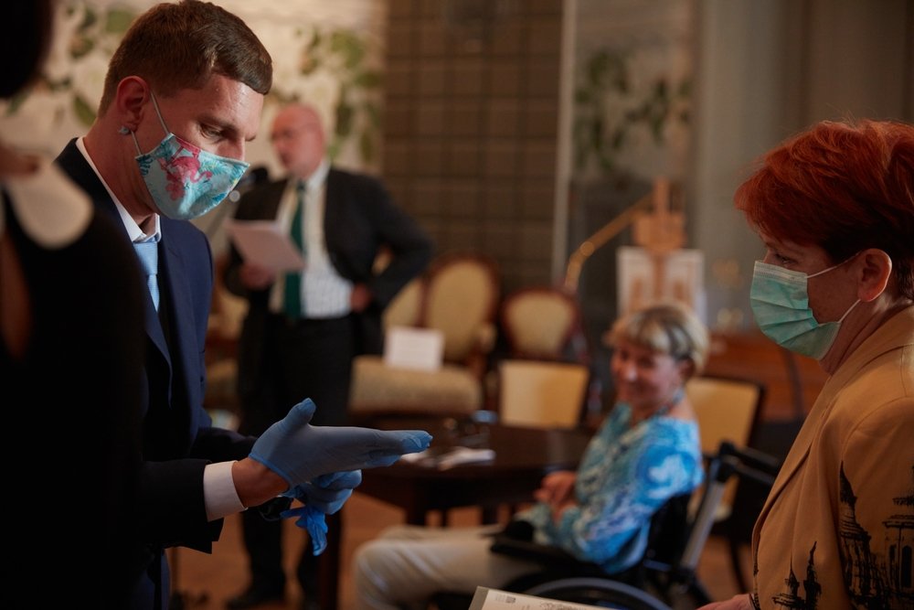 Mężczyzna w garniturze i kolorowej maseczce nakłada niebieskie jednorazowe rękawiczki po lewej stronie kadru, naprzeciw kobiety z krótkimi rudymi włosami i chirurgiczną maseczką, w tle sala z krzesłami i stołem, siedząca osoba na wózku oraz stojący mężczyzna trzymający dokumenty.