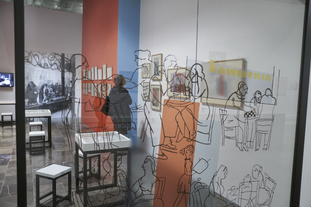 Wnętrze galerii widziane przez przeszkloną ścianę z czarnymi konturowymi rysunkami postaci; za szybą stoi osoba w ciemnym płaszczu oglądająca oprawione prace na ścianie z pionowymi pasami w kolorze niebieskim i pomarańczowym, w przestrzeni ustawione są stoły, gabloty i krzesła, po lewej duża czarno-biała fotografia, po prawej na ścianie widoczny napis "kawiarnia".