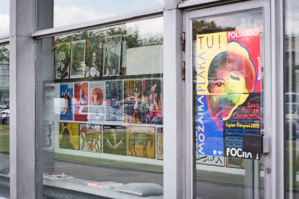 Szklana witryna i drzwi w metalowej ramie, przez szybę widać wewnętrzną ekspozycję wielu mniejszych plakatów ułożonych w siatkę oraz po prawej duży, kolorowy plakat z wyraźnie stylizowaną twarzą i pionowym napisem, na szybie odbicia zaparkowanych samochodów i drzew, a na dolnym parapecie leżą magazyny.