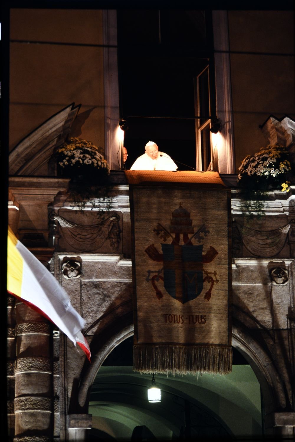 Osoba w białym ubraniu i białej czapce stoi na oświetlonym balkonie za mikrofonem nad dużym ozdobnym sztandarem z herbem i napisem TOTUS TUUS, na kamiennej elewacji z kwiatami po bokach i falującą biało‑żółtą flagą.