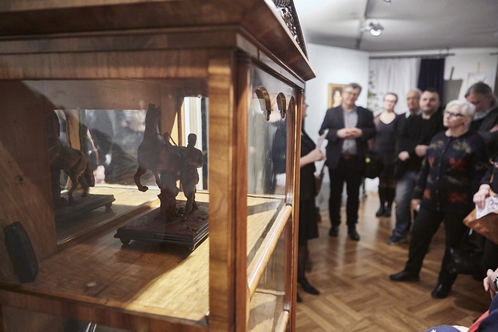Wnętrze z drewnianą, przeszkloną gablotą na pierwszym planie, w której widać małą rzeźbę jeźdźca na koniu i jej odbicie, a w tle rozmyta grupa osób stojąca na drewnianej podłodze przed obrazami na ścianie.