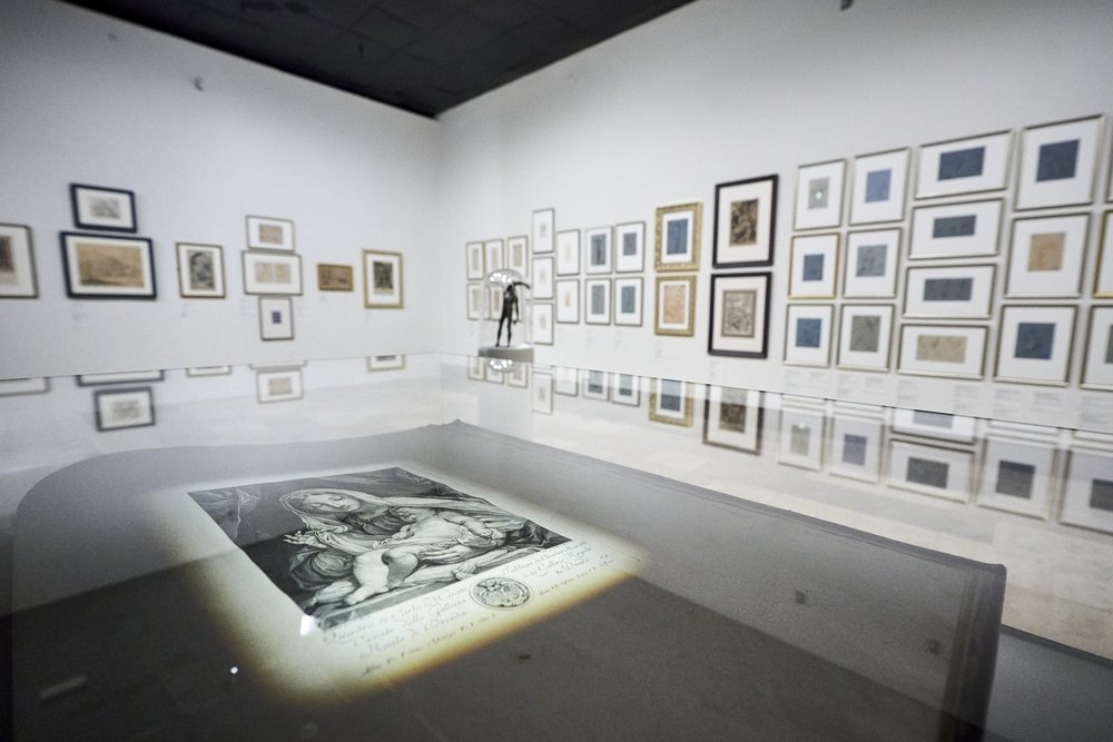 Widok wnętrza galerii z białymi ścianami pokrytymi wieloma oprawionymi rysunkami i grafikami; na pierwszym planie oszklona wystawka z podświetloną czarno-białą grafiką przedstawiającą kobietę trzymającą dziecko, w głębi stoi rzeźba na cokole, a połysk podłogi i szkła odbija obrazy na ścianach.