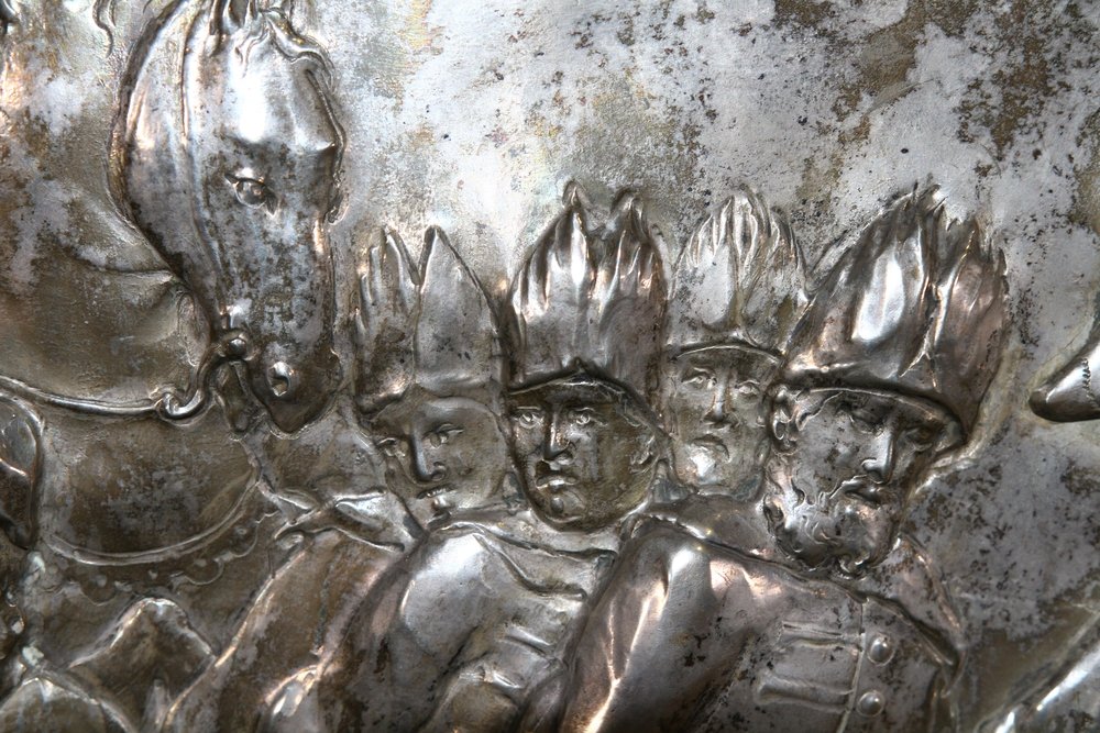 Zbliżenie na wypukły srebrny relief przedstawiający z lewej głowę konia i czterech mężczyzn ustawionych jeden za drugim w wysokich, spiczastych nakryciach głowy, jeden z nich z brodą, z widoczną patyną i zmatowieniem metalu.