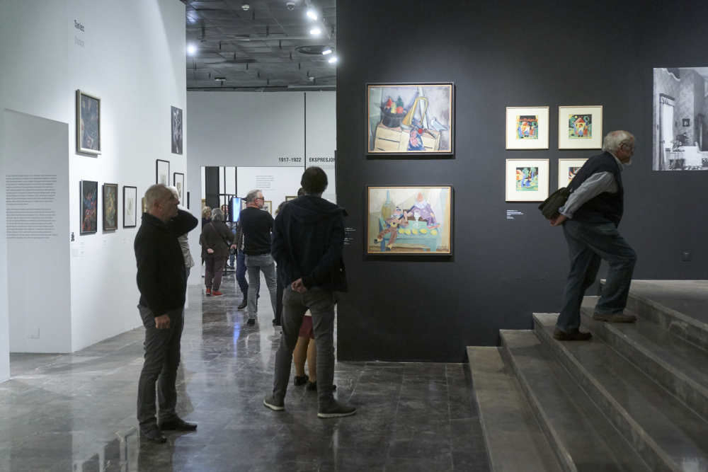 Wnętrze galerii sztuki z kilkoma osobami oglądającymi obrazy; pośrodku ciemnoszara ściana z kilkoma oprawionymi, kolorowymi obrazami, po lewej biała ściana z mniejszymi ramami, błyszcząca posadzka oraz po prawej schody, po których wchodzi mężczyzna, a inni zwiedzający przechadzają się i oglądają eksponaty.