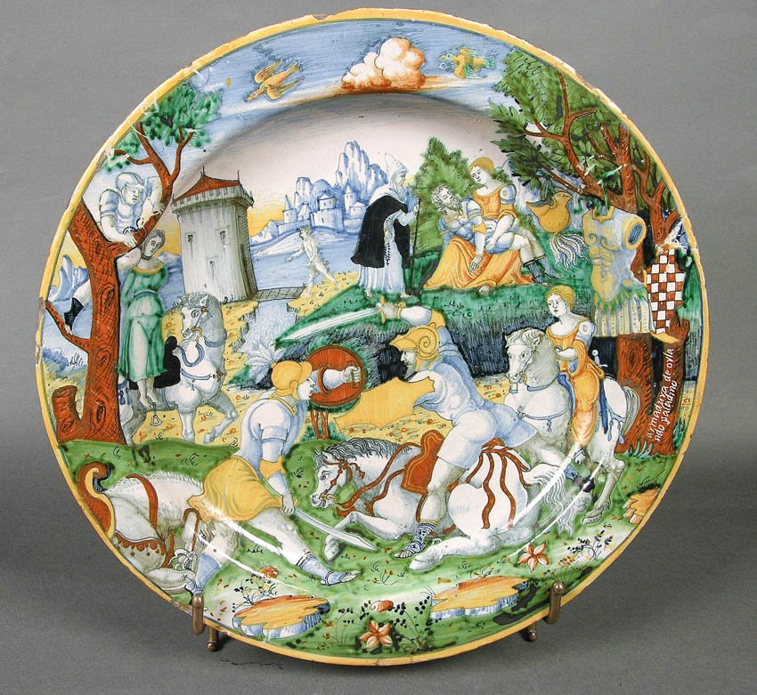 Kolorowy ceramiczny talerz z malowaną sceną bitewną: w centrum rycerze w zbrojach na białych koniach, jeden koń leżący, wokół drzewa i stojące postacie obserwujące, w tle kamienna wieża i skaliste wzgórza, nad nimi niebo z chmurą i lecącymi ptakami, a brzegi talerza ozdobione zielono‑żółto‑niebieskimi motywami roślinnymi.