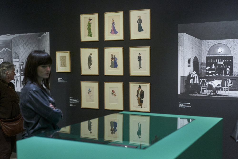 Wnętrze galerii z ciemną ścianą, na której w złotych ramach ułożone w trzy kolumny i trzy wiersze wiszą rysunki postaci; po prawej duża czarno-biała fotografia wnętrza, na pierwszym planie zielona witryna z odblaskiem eksponatów, a przy lewej krawędzi dwie osoby stojące i oglądające wystawę.