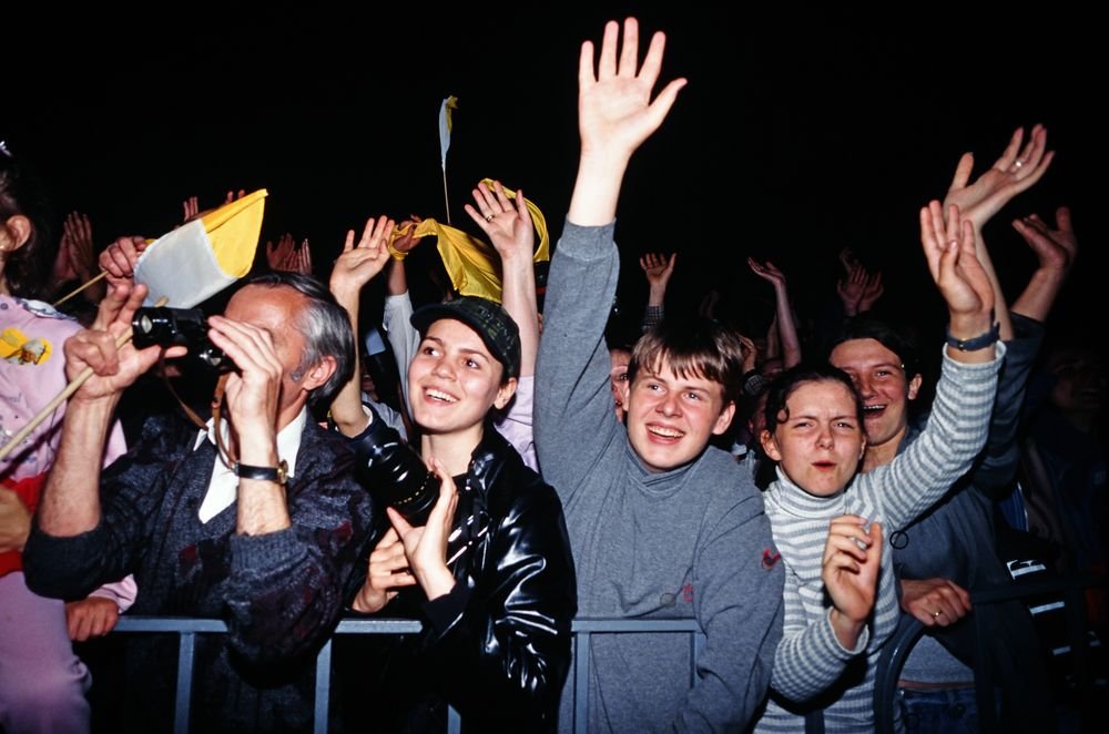 Grupa osób stojąca za barierką na nocnym wydarzeniu z uniesionymi rękami — mężczyzna z lornetką i aparatem, młodzi ludzie (uśmiechnięta dziewczyna w czapce, chłopak w szarej bluzie, dziewczyna w paskowanym swetrze) oraz kilka żółto-białych flag na ciemnym tle.