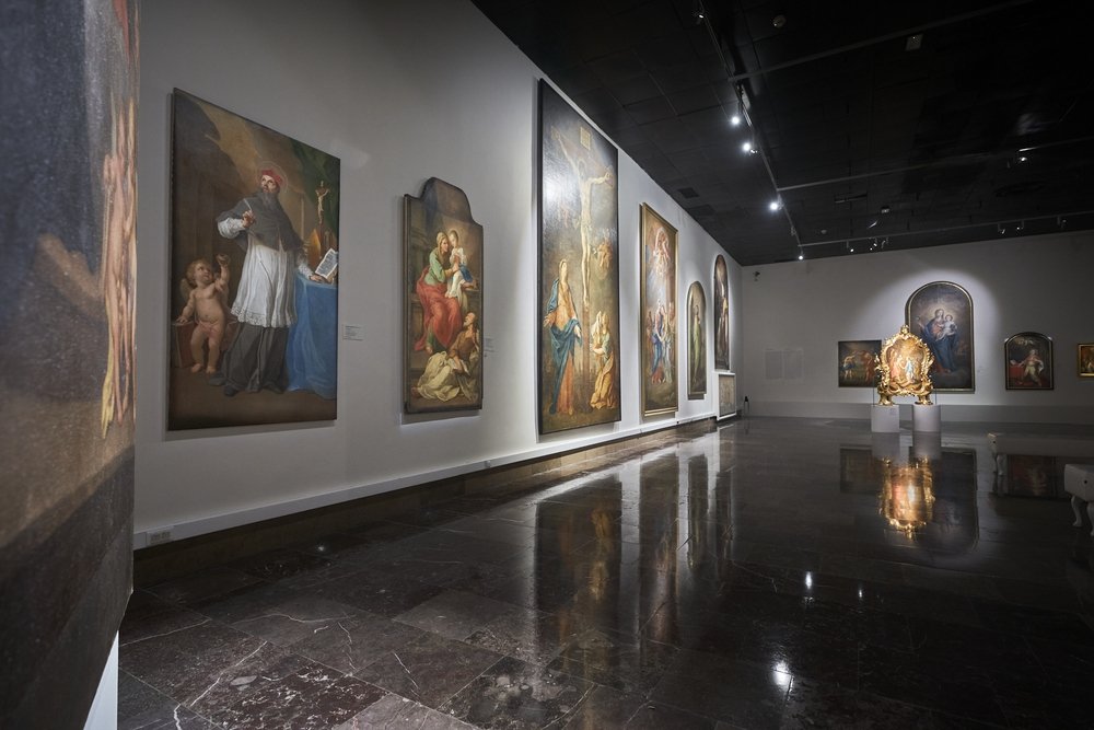 Widok wnętrza galerii sztuki: długa sala z białymi ścianami i ciemną, błyszczącą marmurową podłogą odbijającą światło, przy której wiszą duże obrazy przedstawiające sceny religijne, a po prawej stoi oświetlona złocona rzeźba na postumencie.