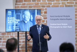 Mężczyzna w ciemnym garniturze i okularach stoi przy mikrofonie na tle ceglanej ściany, za nim wiszą plakaty z czarno-białym portretem i tekstem, a na pierwszym planie widać z tyłu sylwetki kilku słuchaczy.