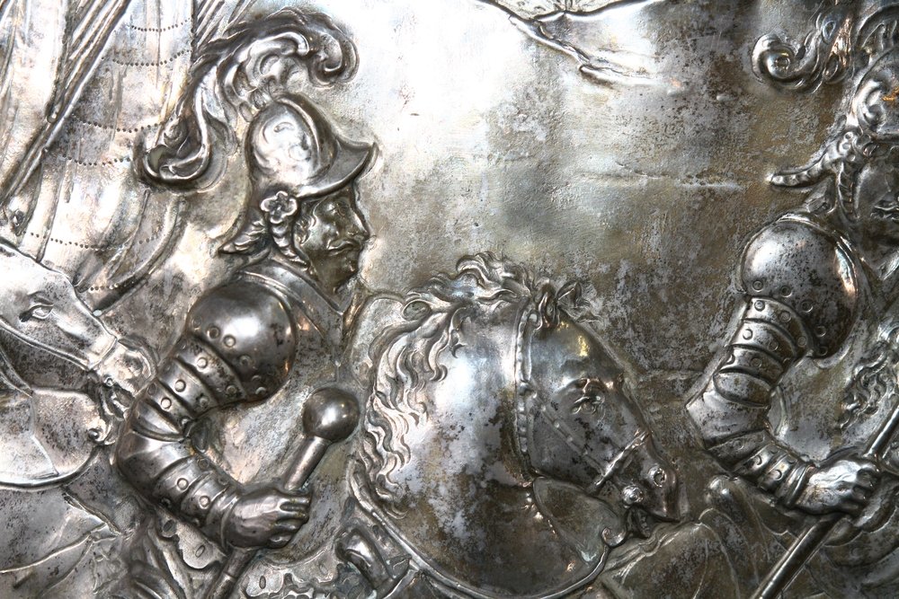 Fragment wypukłej srebrnej płaskorzeźby ukazujący postać w zbroi z ozdobnym hełmem i berłem oraz głowę osiodłanego konia, z widocznymi detalami pancerza i fragmentem drugiej uzbrojonej postaci po prawej.
