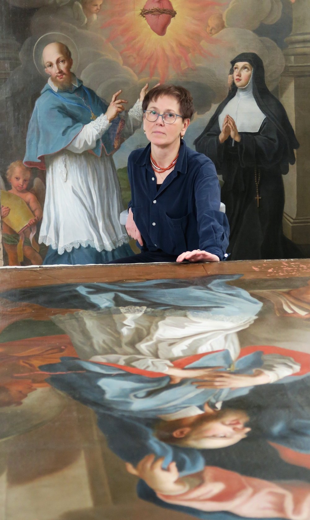 Kobieta w krótkich włosach i okularach, w granatowej koszuli i czerwonym naszyjniku, pochylona nad dużym odwróconym obrazem leżącym na stole; w tle rozległe religijne malowidło z mężczyzną w niebieskiej szacie i aureolą, klęczącą zakonnicą w czarnym habicie, dwoma cherubinami oraz świecącym sercem w koronie cierniowej.