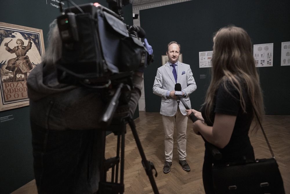 Operator kamery po lewej stoi za dużą kamerą na statywie, przed nim stoi mężczyzna w jasnoszarej marynarce, koszuli i fioletowym krawacie trzymający mikrofon w sali wystawowej z ciemnozielonymi ścianami i plakatami, a z prawej stoi kobieta z długimi włosami wyciągająca drugi mikrofon; na podłodze widoczny drewniany parkiet.