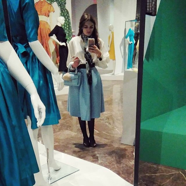 Osoba robiąca selfie w lustrze przed ekspozycją sukien; na pierwszym planie manekiny w turkusowych sukienkach po lewej, w tle manekiny w pomarańczowej, zielonej w kropki, czarnej i żółtej stylizacji; osoba ubrana w białą bluzkę, jasnoniebieską rozkloszowaną spódnicę, czarne rajstopy i czarne buty, z wzorzystą apaszką i jasnoniebieską torebką, stojąca na marmurowej posadzce obok przeszklonych gablot.
