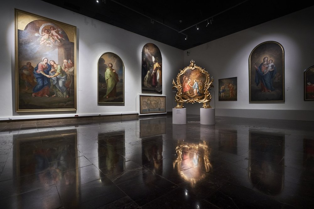 Wnętrze galerii z białymi ścianami i ciemną, błyszczącą posadzką odbijającą światło, na których wiszą duże obrazy olejne o prostokątnych i zaokrąglonych górnych krawędziach przedstawiające postacie i sceny, a pośrodku na dwóch białych cokółach stoi bogato złocona, rzeźbiona rama z malowidłem, doświetlone reflektorami sufitowymi.