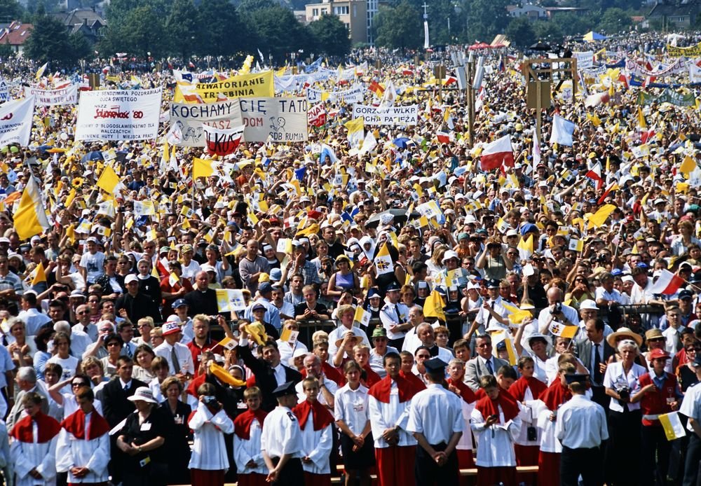 Ogromny plenerowy tłum ludzi, z przodu osoby w białych i czerwonych tunikach, za nimi gęsto zgromadzeni uczestnicy trzymający liczne żółto‑białe i biało‑czerwone flagi oraz duże banery z polskimi napisami (m.in. "KOCHAMY CIĘ TATO"), w tle drzewa i zabudowania.