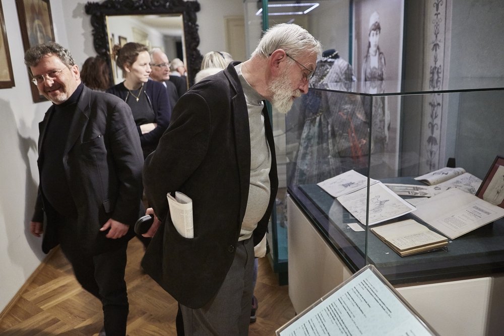 Grupa zwiedzających w muzeum, na pierwszym planie starszy mężczyzna z białą brodą pochylony nad przeszkloną gablotą z książkami i rysunkami, obok uśmiechnięty mężczyzna w ciemnym ubraniu, w tle inne osoby, duże lustro i wystawiona historyczna suknia.