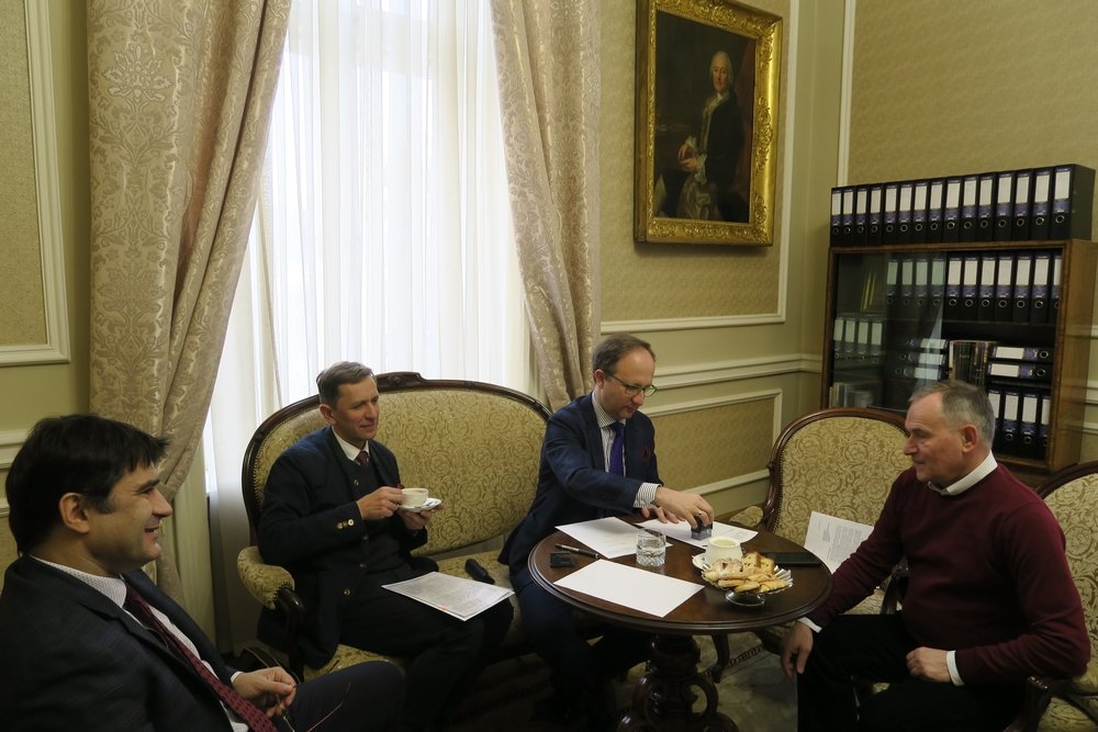Czterech mężczyzn siedzi przy małym okrągłym drewnianym stoliku w eleganckim, tapicerowanym pokoju z wysokim oknem i zasłonami; na stole leżą dokumenty, filiżanki i talerz z przekąskami, jeden mężczyzna trzyma filiżankę, inny sięga po przedmiot, a na ścianie wisi oprawiony portret obok regału z segregatorami.