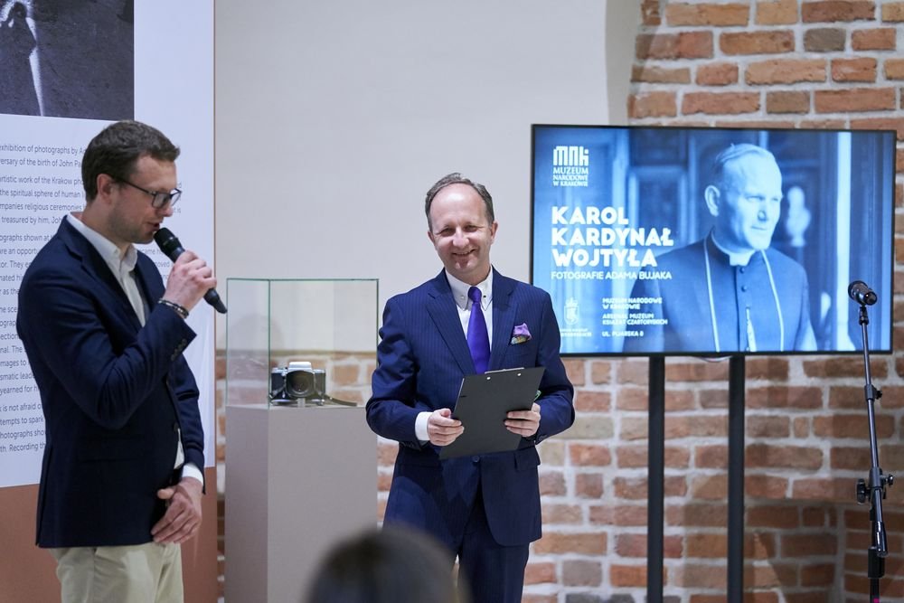 Dwóch mężczyzn w eleganckich garniturach stoi przy ekspozycji w sali z ceglaną ścianą; mężczyzna po lewej w okularach trzyma mikrofon, mężczyzna po prawej w granatowym garniturze z fioletowym krawatem i poszetką uśmiecha się i trzyma czarną teczkę, obok na cokole w szklanej gablocie leży aparat fotograficzny, a za nimi widoczny jest ekran z czarno-białym zdjęciem duchownego i napisem "KAROL KARDYNAŁ WOJTYŁA".