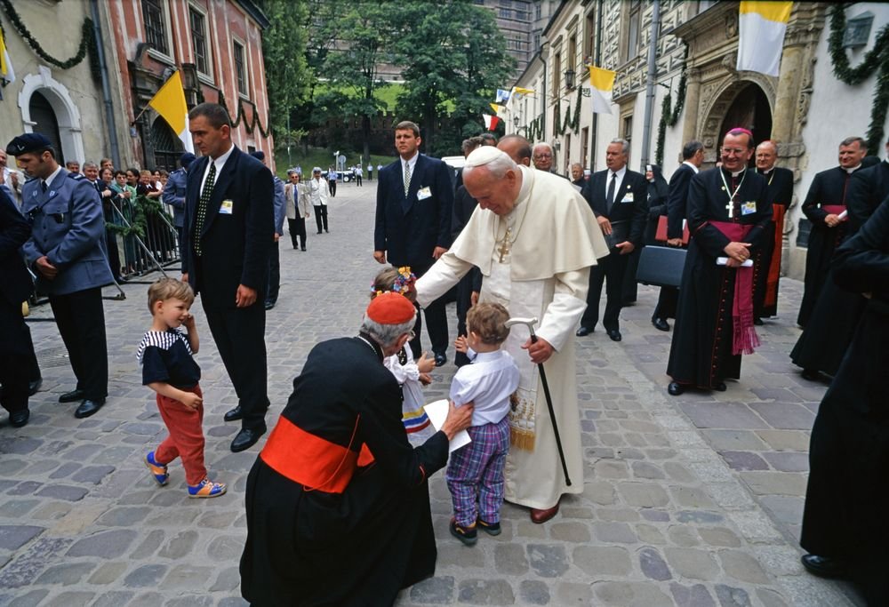 Na brukowanej ulicy grupka duchownych i ochrony otacza starszego mężczyznę w białym stroju religijnym i białej piusce, który pochyla się i dotyka głowy małego chłopca, obok klęczy duchowny w czarnym ubraniu z czerwoną szarfą i piuską, a wokół stoją inni księża, ochroniarze i widzowie przy udekorowanych budynkach.