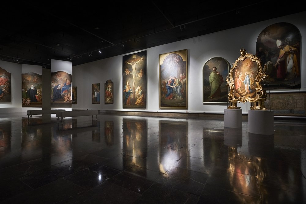 Wnętrze przyciemnionej galerii z lśniącą, odbijającą światło posadzką; na białych ścianach rozmieszczone duże obrazy przedstawiające sceny religijne, pośrodku kilka ławek, a po prawej złocona, bogato zdobiona rama na cokole.