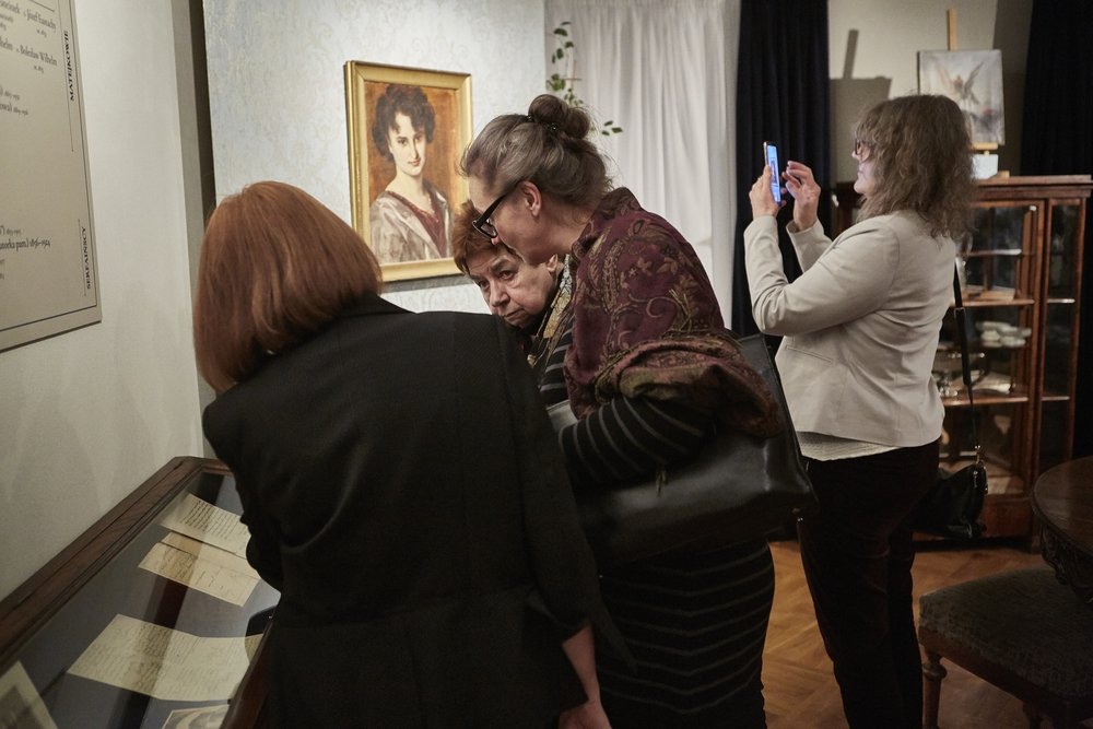 Cztery kobiety w sali wystawowej przy przeszklonej gablocie z dokumentami — trzy pochylone nad ekspozycją, czwarta w jasnej marynarce robi zdjęcie telefonem; na ścianie nad gablotą oprawiony portret, w tle drewniana witryna z naczyniami i meble.