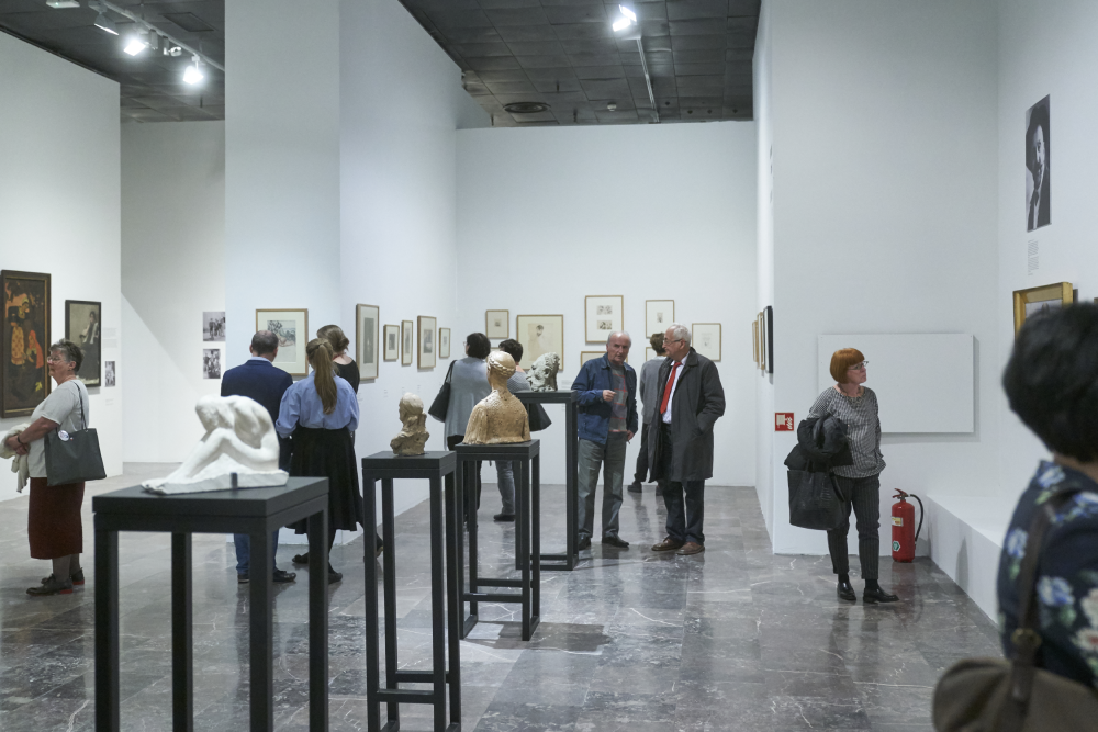 Wnętrze galerii z białymi ścianami i ciemną marmurową podłogą, na pierwszym planie kilka rzeźb ustawionych na wysokich czarnych postumentach, w tle grupy osób oglądające ramy i grafiki wiszące na ścianach, sufit z reflektorami oraz widoczna gaśnica przy prawej ścianie.