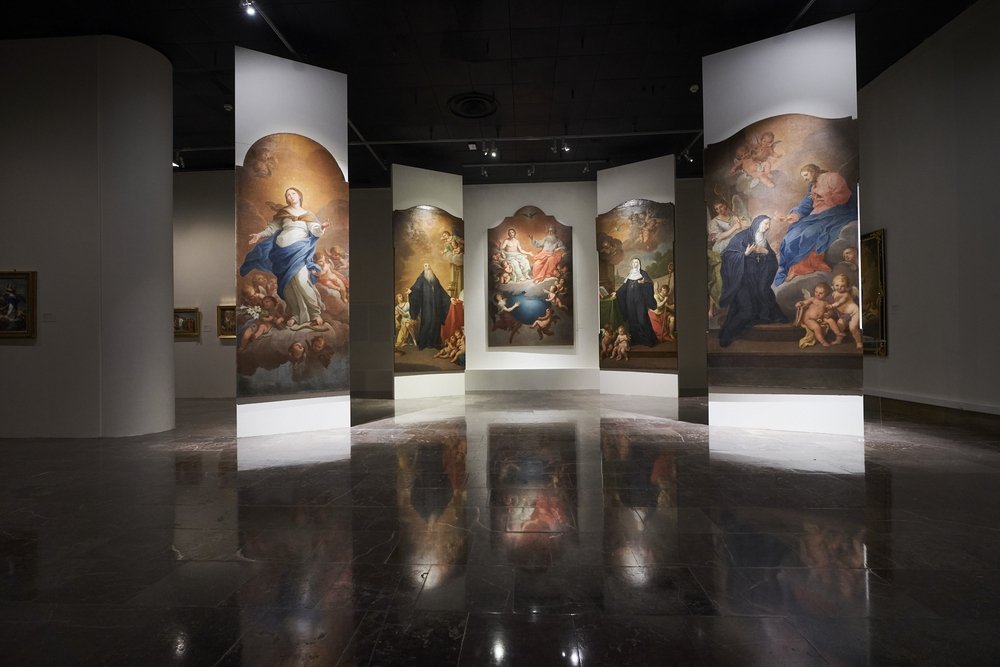 Wnętrze ciemnej galerii z lśniącą marmurową podłogą i pięcioma wysokimi, wolnostojącymi białymi panelami z dużymi klasycznymi obrazami przedstawiającymi postaci, anioły i putta, których odbicia widać na posadzce.