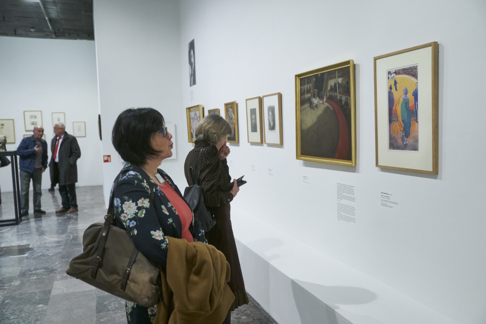 Wnętrze galerii sztuki z białą ścianą, na której w szeregu wiszą oprawione obrazy i grafiki (po prawej wyraźny kolorowy obraz w jasnej ramie i obraz w złotej ramie); na pierwszym planie dwie kobiety oglądają eksponaty — jedna w kwiecistej marynarce z torebką i płaszczem, druga w ciemnym płaszczu trzyma telefon i czyta podpisy; w tle dwóch mężczyzn rozmawia, marmurowa posadzka i tabliczki z opisami przy obrazach.