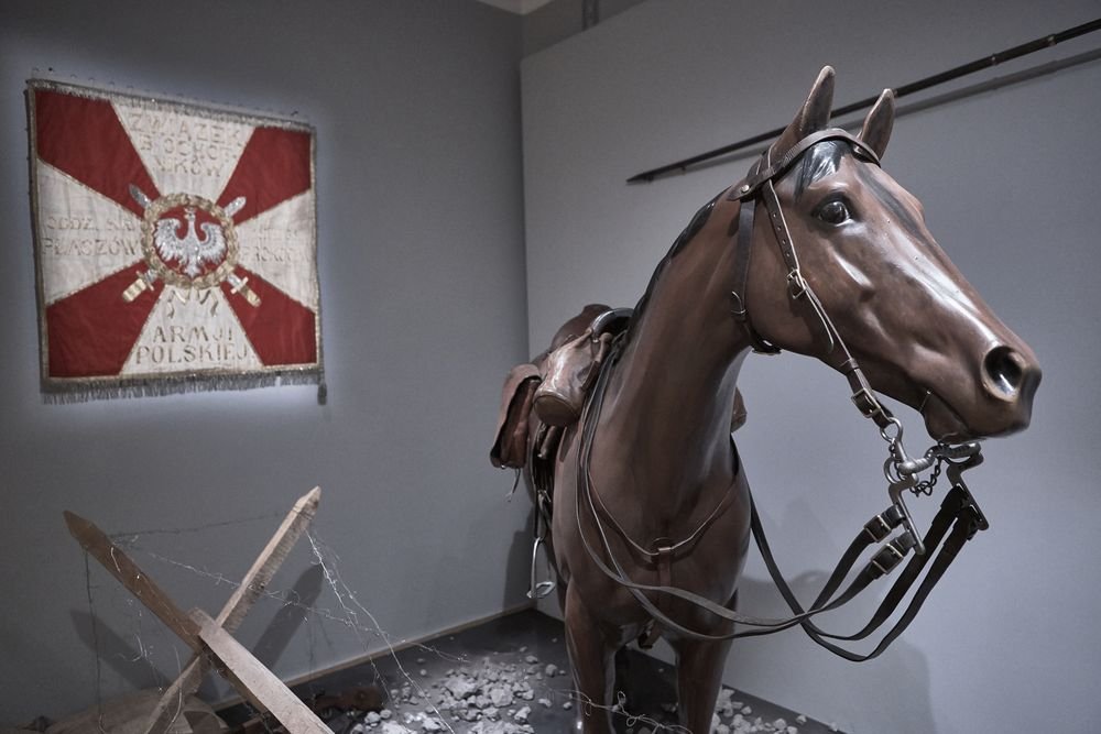 Model konia w uprzęży i siodle zajmający prawą stronę zdjęcia, po lewej wiszący czerwono-biały sztandar z orłem i fragmentem napisu, u dołu drewniane zapory i rozciągnięty drut kolczasty na podłodze.