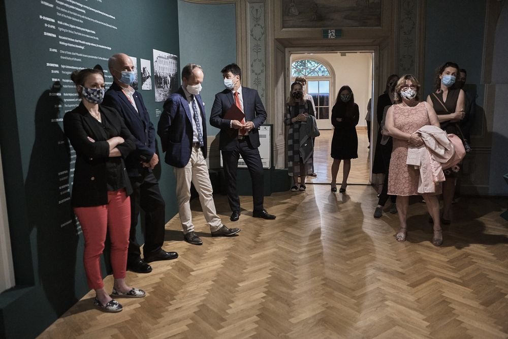 Kilka dorosłych osób w maseczkach stojących w grupach po lewej i prawej stronie sali wystawowej przy ciemnozielonej ścianie z tekstem i zdjęciami, na jodełkowym drewnianym parkiecie, z otwartymi drzwiami i oknem widocznym w głębi.