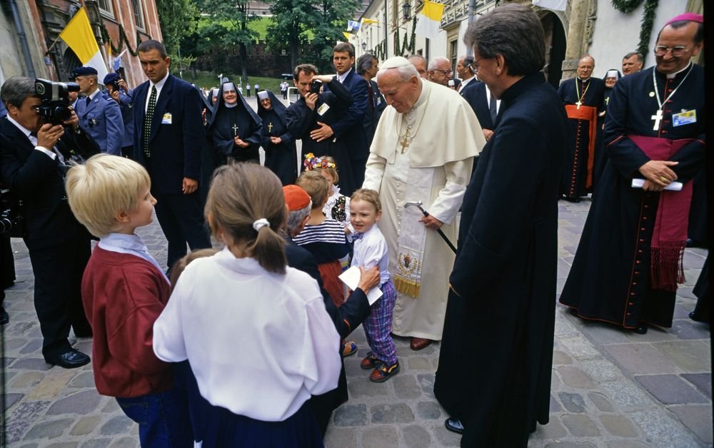 Na brukowanym placu przed historycznymi budynkami stoi mężczyzna w białych szatach liturgicznych trzymający pastorał, otoczony duchownymi i biskupami, za nimi kilka zakonnic, ochroniarze i fotografowie, a z przodu grupa dzieci rozmawiająca z dorosłymi.