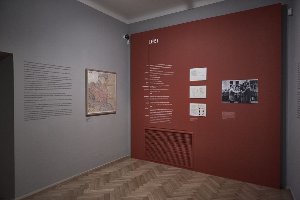 Pomieszczenie wystawowe z szarą i bordową ścianą; na bordowej ścianie duży napis „1921”, oś czasu z tekstem, trzy dokumenty i czarno-biała fotografia, po lewej na szarej ścianie opisowa plansza i oprawiona mapa, na podłodze jodełkowe deskowanie oraz kamera w górnym narożniku.