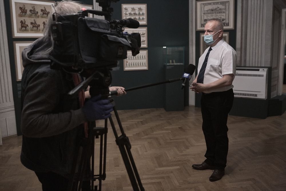 Operator kamery z profesjonalnym sprzętem na statywie filmuje stojącego na drewnianej podłodze mężczyznę w krótkim rękawie, krawacie i jednorazowej maseczce, a do niego wyciągnięty jest wysięgnik z mikrofonem studyjnym z widoczną flagą, w tle ciemnozielona ściana galerii z oprawionymi grafikami i gablotą.