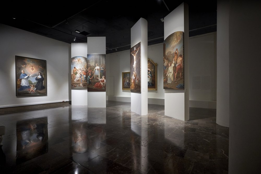 Wnętrze galerii z kilkoma wysokimi białymi wolnostojącymi panelami i ścianami, na których wiszą duże klasyczne obrazy o tematyce religijnej (w tym centralnie widoczny obraz ukrzyżowania), ciemną błyszczącą marmurową posadzką odbijającą prace oraz punktowym oświetleniem sufitowym.