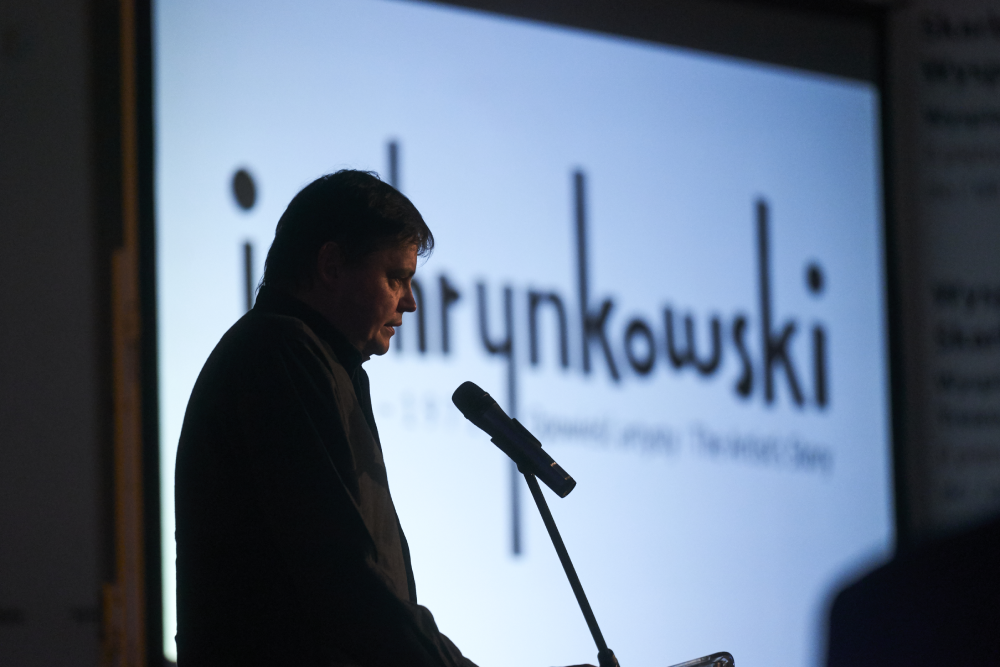 Sylwetka mężczyzny w ciemnym ubraniu stojącego przy mikrofonie na statywie na tle dużego, jasnego ekranu projekcyjnego z rozmytym czarnym napisem.