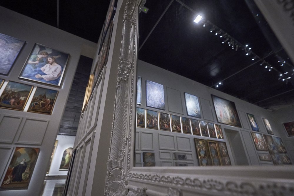 Wnętrze galerii z licznymi oprawionymi obrazami na ścianach, w pierwszym planie ozdobna biała rama lustra odbijająca rząd obrazów i sufit z punktowymi reflektorami.